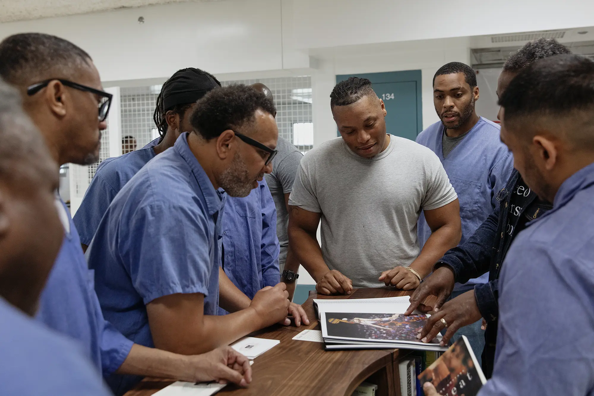 Men clad in blue gather around a wooden Freedom Library bookcase discussing a copy of Reginald Dwayne Betts and Titus Kaphar’s Redaction book.