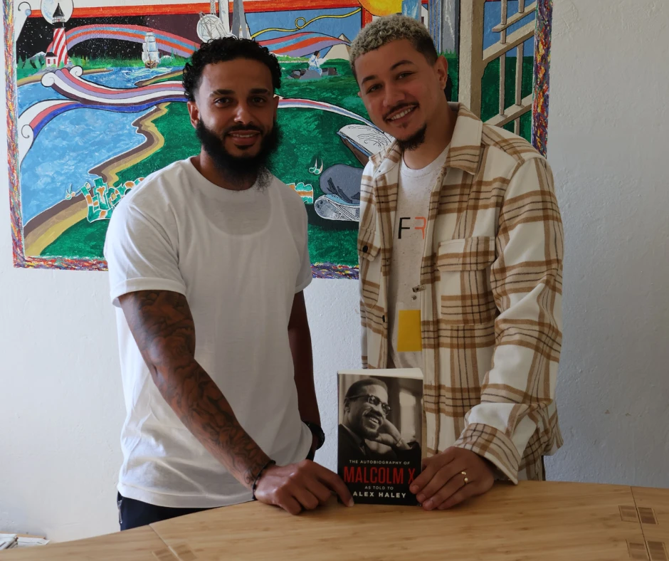 Freedom Library Patron Hector stands with Freedom Reads Library Coordinator David Perez DeHoyos at MCI-Norfolk holding a Malcolm X book standing behind the Freedom Library.