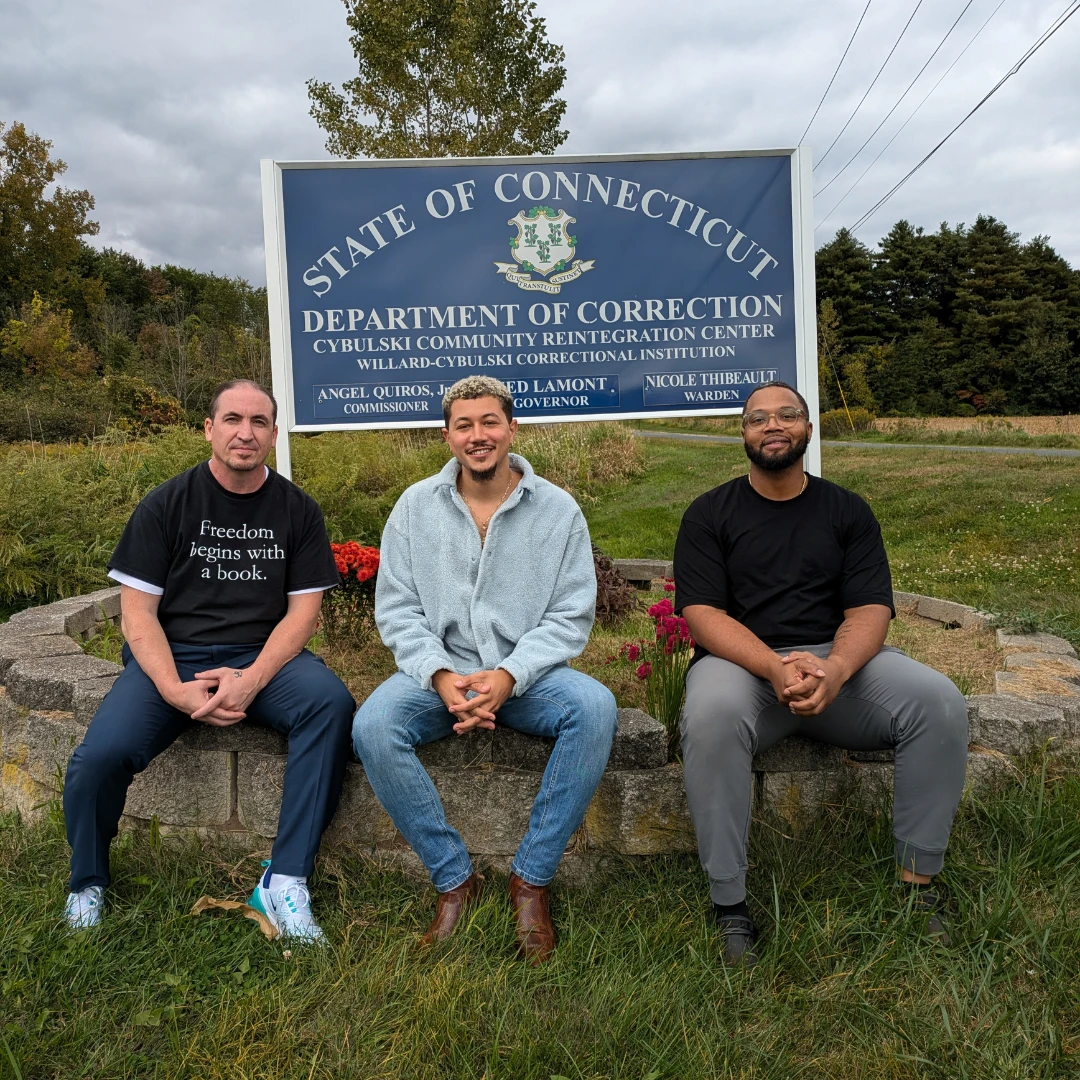 Freedom Reads Communications Manager Steven Parkhurst (left), Freedom Reads Library Coordinator David Perez Jr (center), and Freedom Reads Library Production Assistant Michael Byrd (right) outside of Cybulski Community Reintegration Center. Freedom Reads Communications Manager Steven Parkhurst (left), Freedom Reads Library Coordinator David Perez Jr (center), and Freedom Reads Library Production Assistant Michael Byrd (right) sit outside of a sign for the Cybulski Community Reintegration Center.