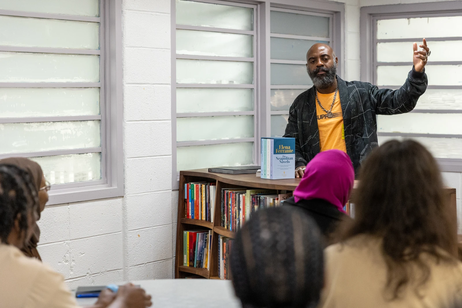 Dwayne speaks, arm raised, in front of a freedom library bookcase to a group of women in khaki prison uniforms.
